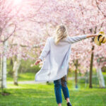 Blonde woman walking through a park of cherry trees waving her hat in happiness.
